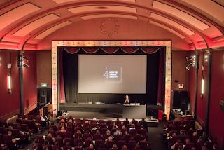 Looking from the balcony at the audience and the stage during a talk. The interior of the cinema is plush with a retro vintage vibe.
