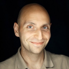 A shaven-headed bright-eyed man smirking against a dark background.