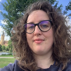 A curly-haired woman with glasses taking a selfie outdoors.